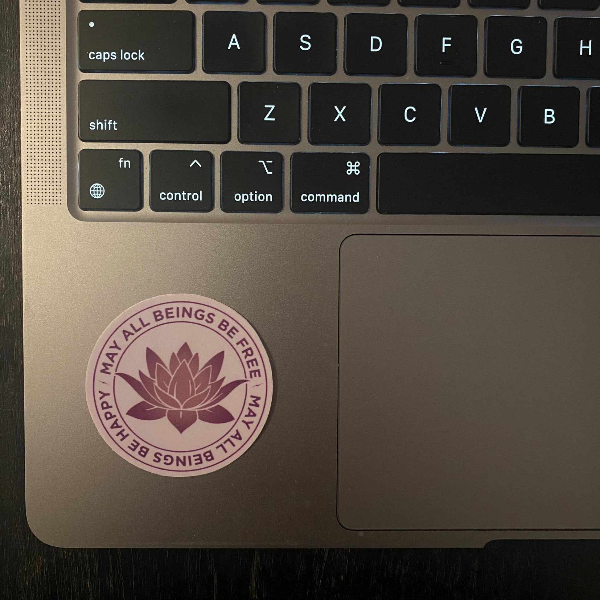 Laptop with a 2" purple sticker with a lotus design and the text, may all beings be free may all beings be happy.