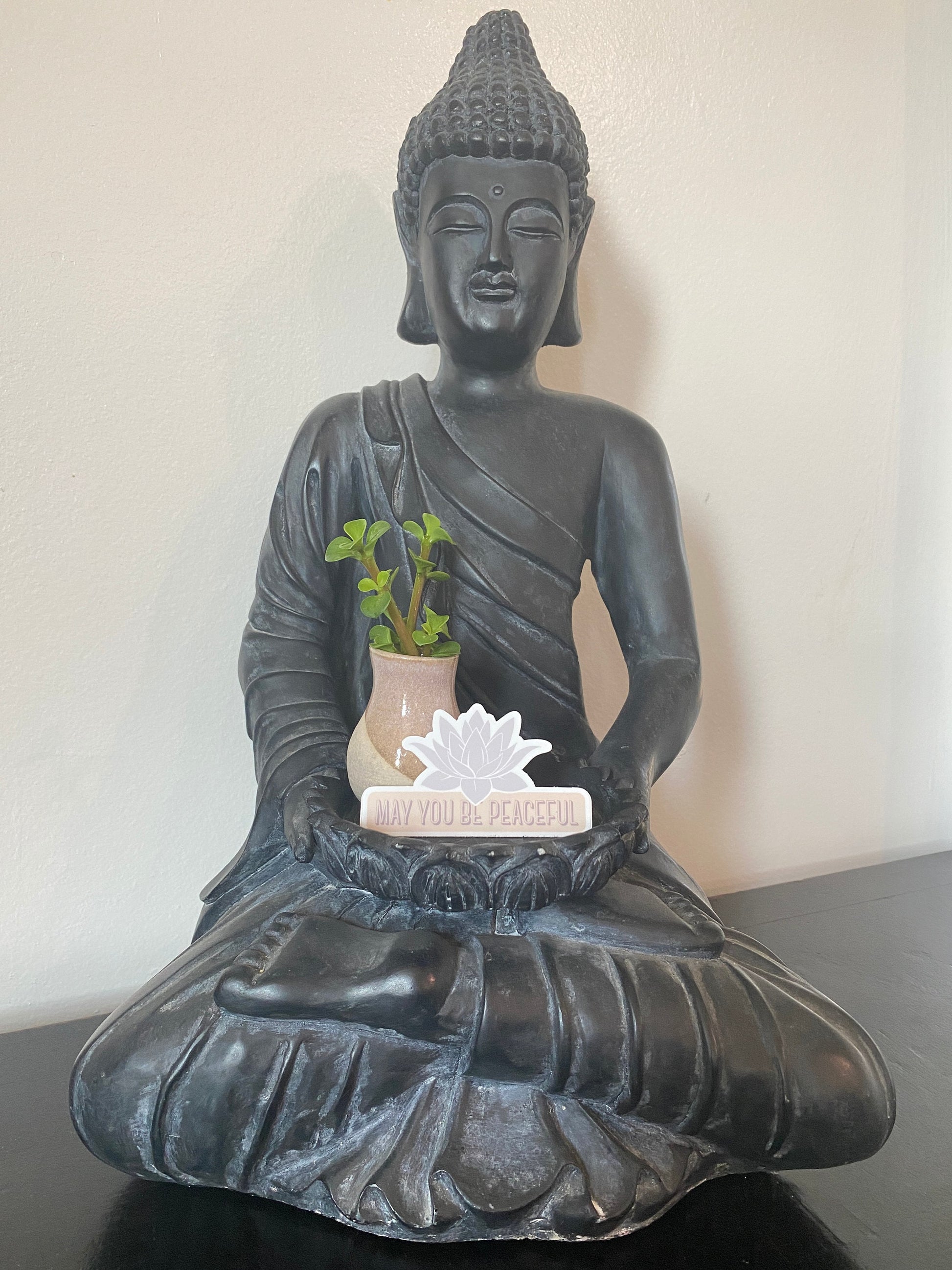 Black Buddha statue holding a plant and a pale lavender lotus design die cut vinyl sticker with the text, may you be peaceful. 