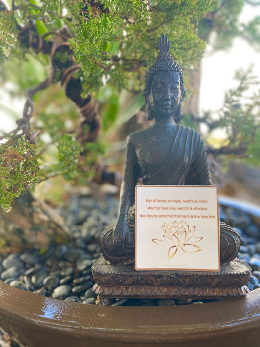 Buddha statue with a Buddhist prayer, 3" square Buddhist sticker with a lotus flower design and text, 'May all beings be happy, healthy and whole. May they have love, warmth and affection. May they be protected from harm and free from fear.' Sticker in front of a bonsai tree.