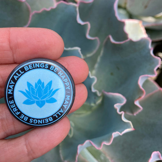 Hand holding a circular blue and black 1.25" button with a lotus flower design and text, may all beings be happy may all beings be free, against a green leafy background.