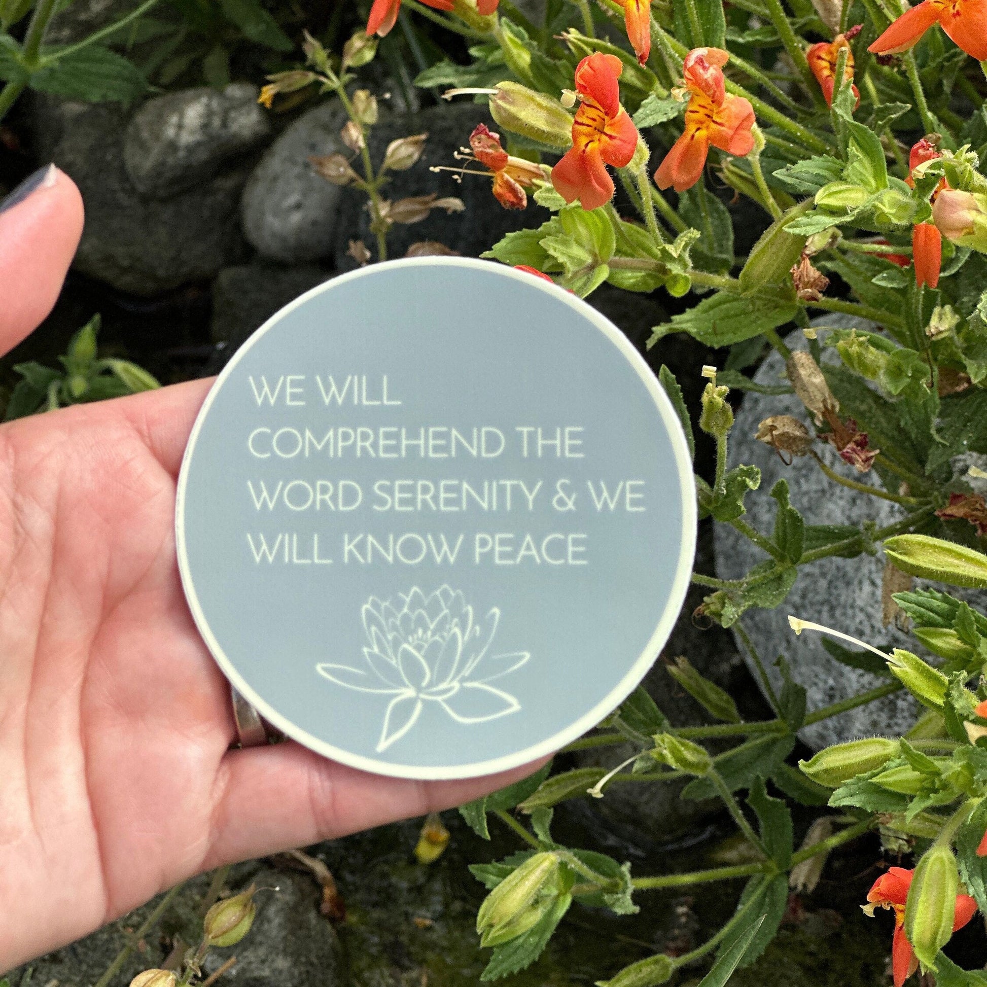 Round teal sticker with, WE WILL COMPREHEND THE WORD SERENITY AND WE WILL KNOW PEACE, quote held over a natural background with flowers and stones.