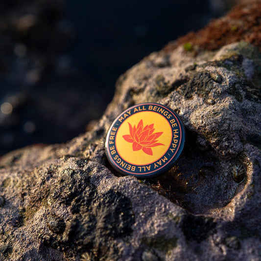 1.25" Orange round pin back button with a buddhist lotus flower and the text 'may all beings be happy may all beings be free' on a textured rock surface.