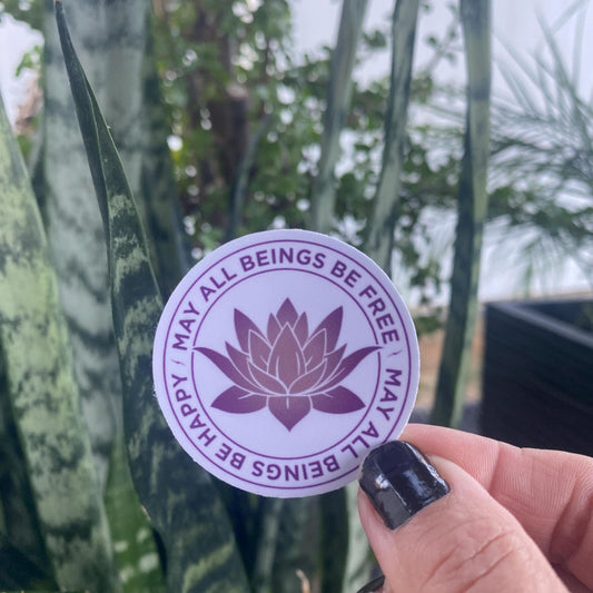 2" purple sticker with a lotus design and the text, may all beings be free may all beings be happy, it is being held in a hand with plants in the background.