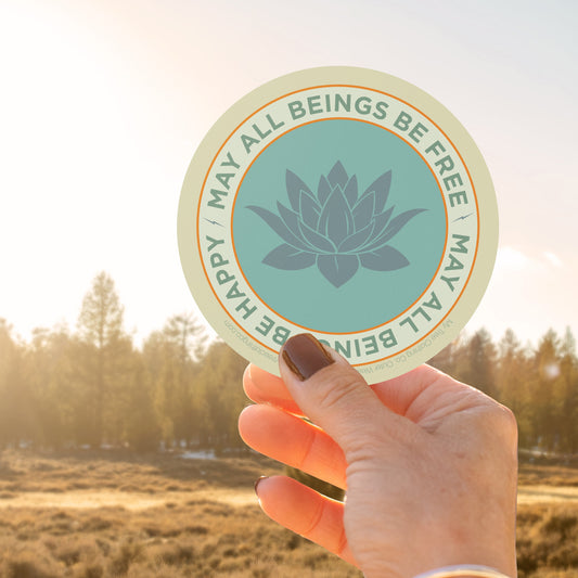 Hand holding a circular sticker with a lotus flower design and text , may all beings be free may all beings be happy, against a natural background.