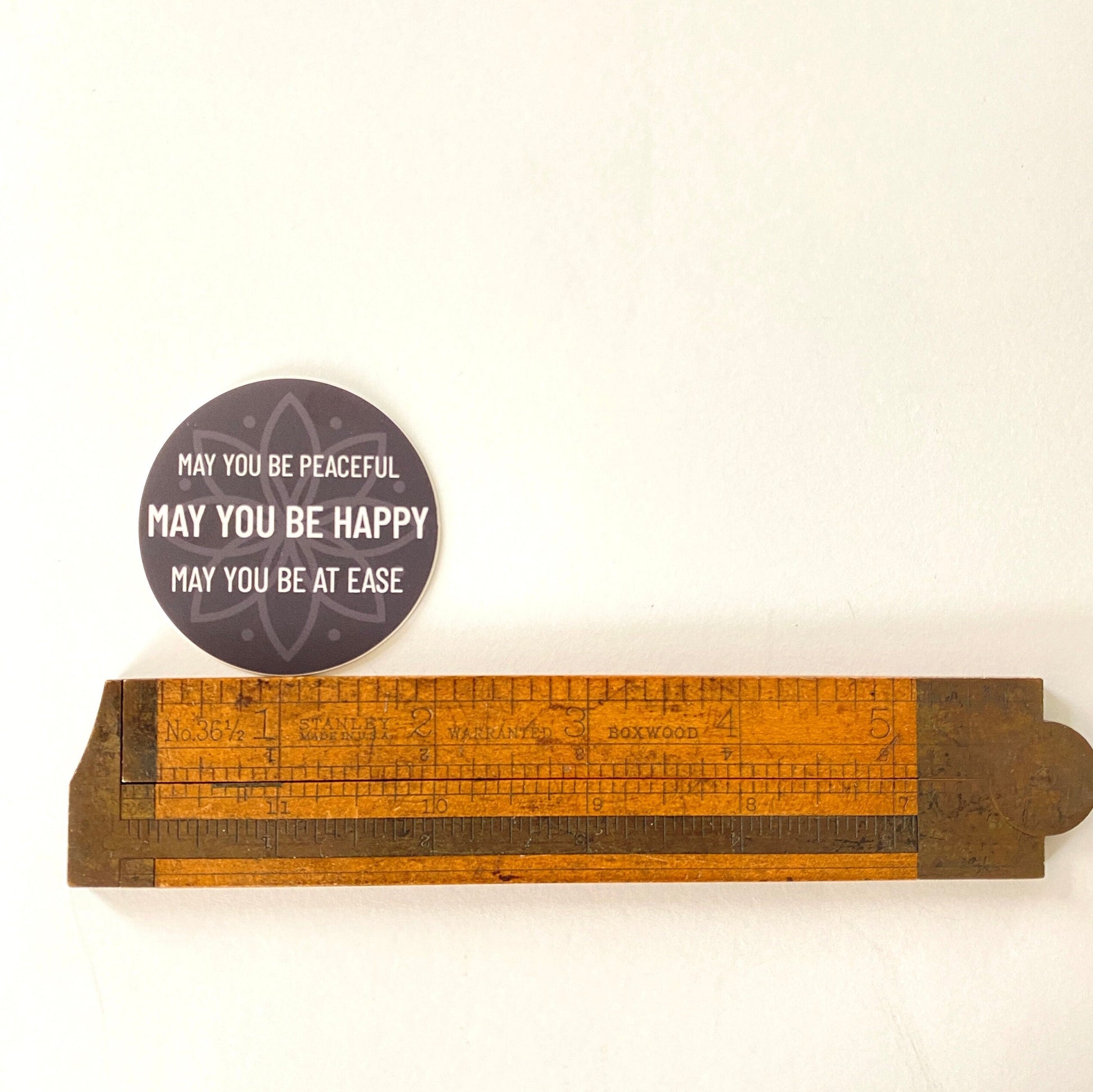 Wooden ruler with a 2" circular sticker with text, may you be peaceful may you be happy may you be at ease, on a white background.