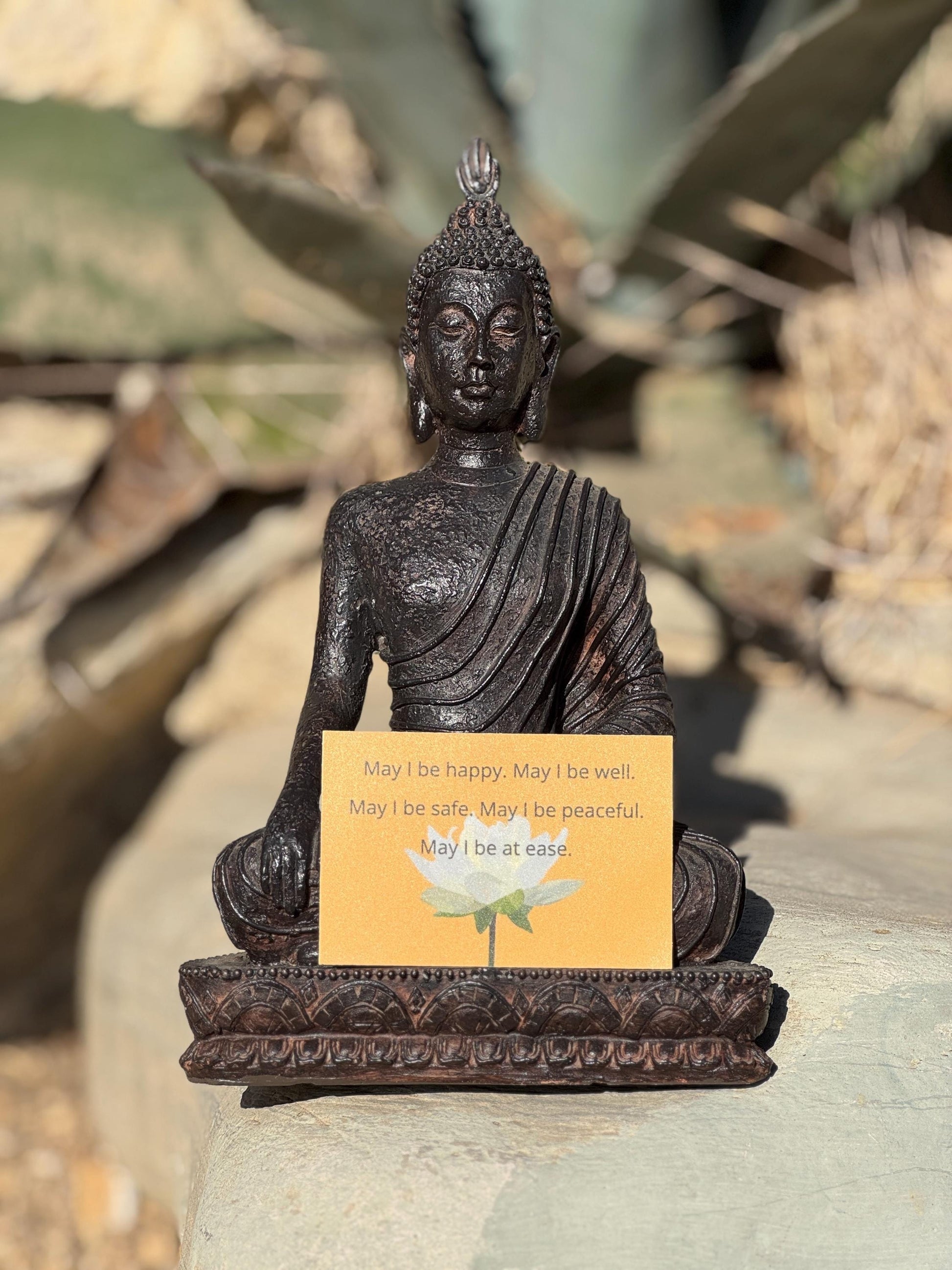 Buddha statue with a rectangular yellow sticker with a lotus and the words, may i be happy may i be well may i be safe may i be peaceful, on a rock with plants in the background.