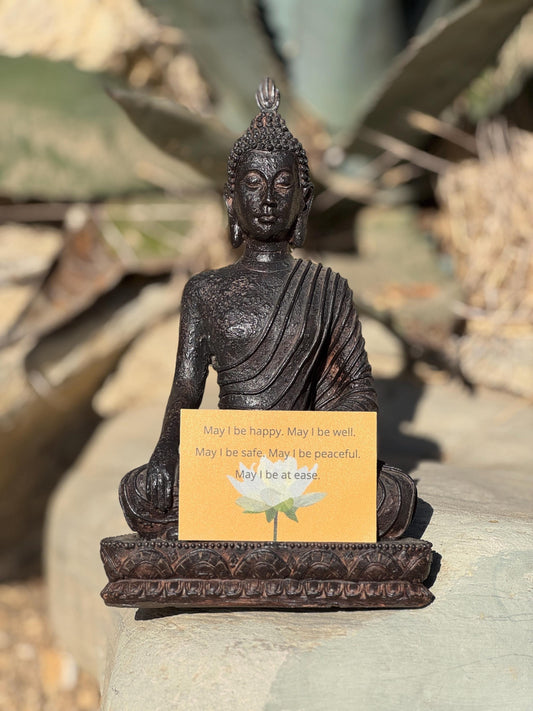 Buddha statue with a rectangular yellow sticker with a lotus and the words, may i be happy may i be well may i be safe may i be peaceful, on a rock with plants in the background.