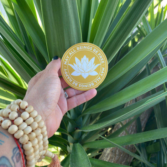 Hand holding a 3" gold and white sticker with a lotus flower design and text, may all beings be free may all beings be happy, against a green leafy background.