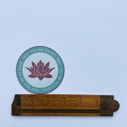 Circular sticker with lotus flower design and text that reads, "may you be happy may you be peaceful may you be at ease" on wooden ruler for scale.