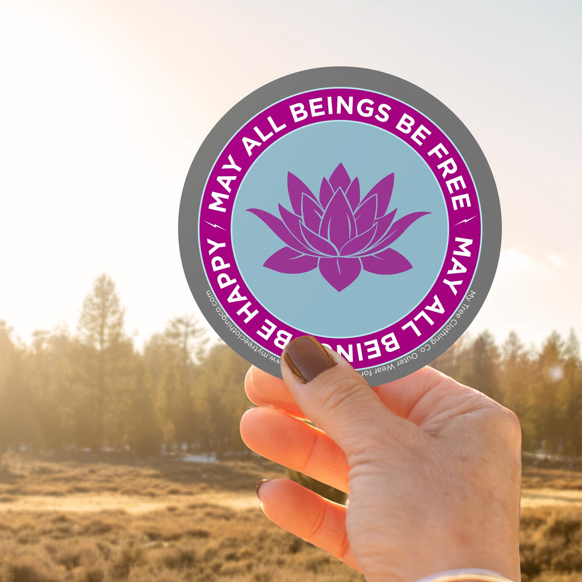Hand holding a 4" circular Buddhist sticker with a purple lotus flower and text, may all beings be free may all beings be happy, against a blurred natural background.