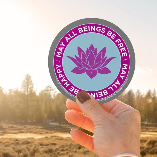 Hand holding a 4" circular Buddhist sticker with a purple lotus flower and text, may all beings be free may all beings be happy, against a blurred natural background.