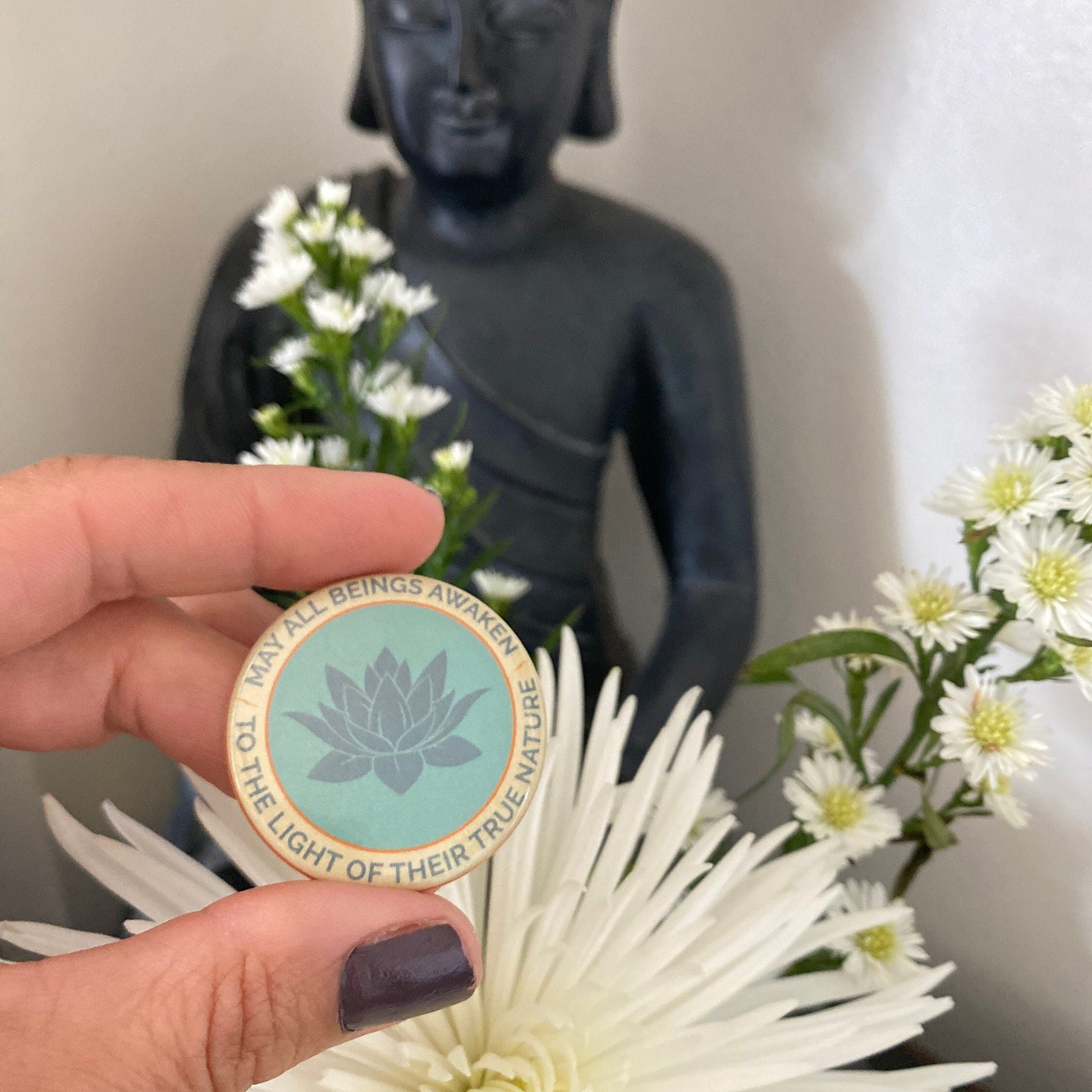 Hand holding a 1.5" button with the mantra that reads, may all beings awaken to the light of their true nature, with a lotus design in front of a Buddha statue and flowers.