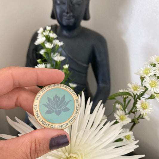 Hand holding a 1.5" button with the mantra that reads, may all beings awaken to the light of their true nature, with a lotus design in front of a Buddha statue and flowers.