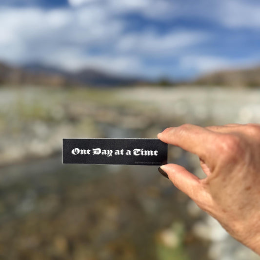 Hand holding a black and white, old english font 'One Day at a Time' sticker with a blurred natural background.