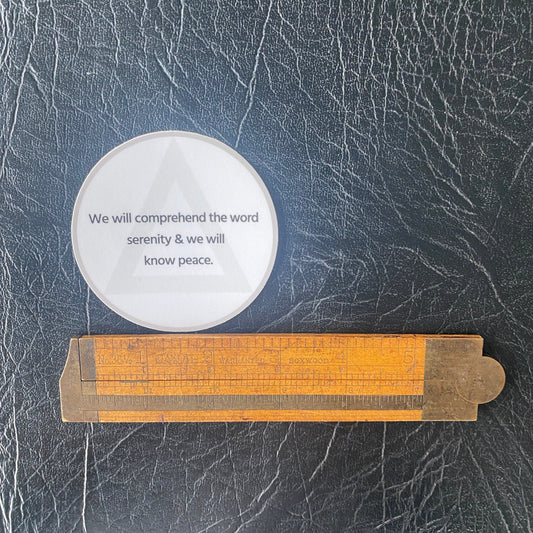 Three inch round white sticker with AA circle and triangle design and the quote, we will comprehend the word serenity and we will know peace. It is being measured by an antique wooden ruler.