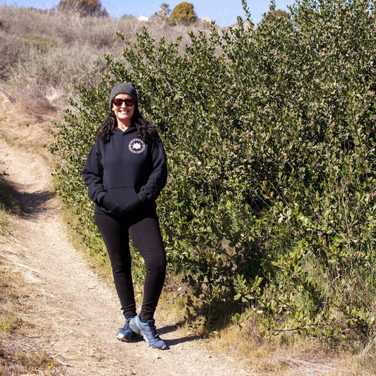 Person wearing Black hoodie with a white lotus and the mettā prayer, 'may you be peaceful may you be happy may you be free.' with leggings standing on a trail with green bushes and trees in the background.