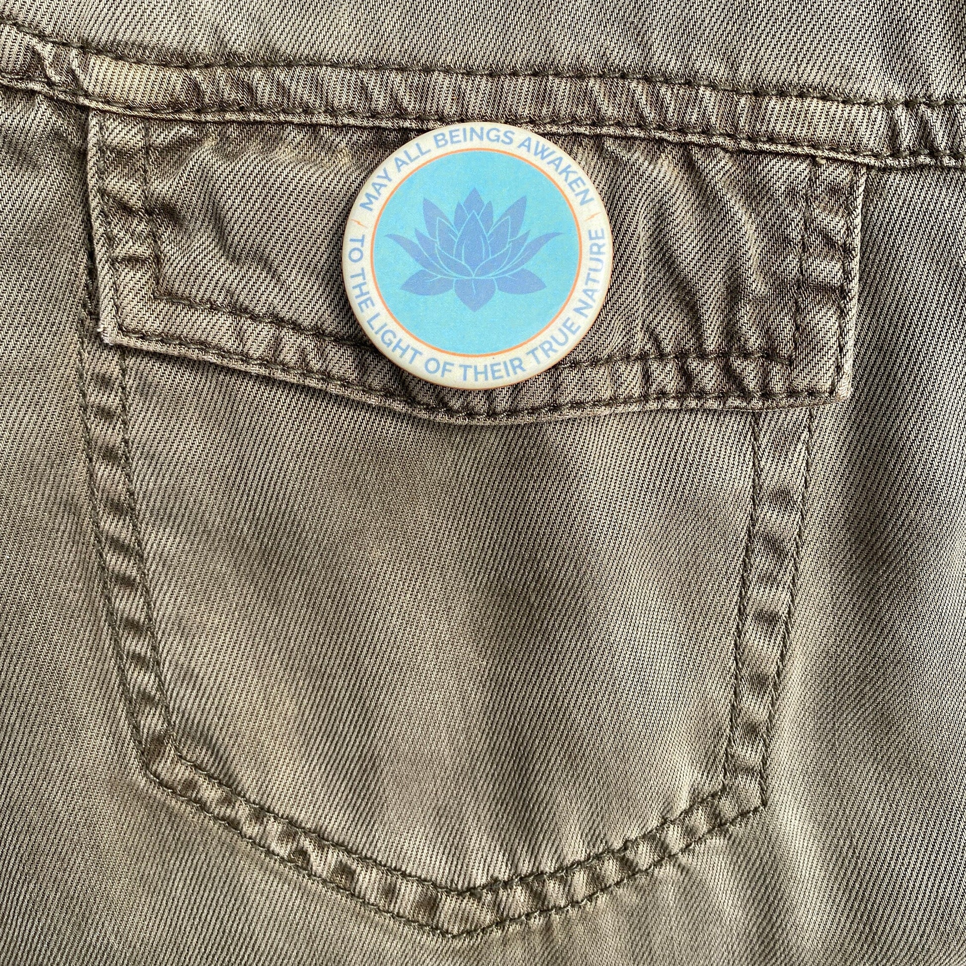 1.5" button with the mantra that reads, may all beings awaken to the light of their true nature, with a lotus design on a gray denim pocket.