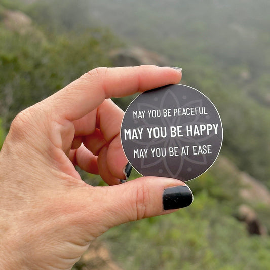 Hand holding a 2" circular sticker with text, may you be peaceful may you be happy may you be at ease, against a blurred natural background