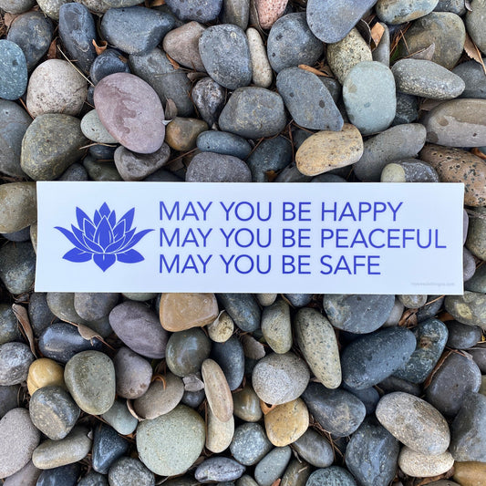 White and blue buddhist prayer bumper sticker that reads, MAY YOU BE HAPPY MAY YOU BE PEACEFUL MAY YOU BE SAFE. On a bed or stones.