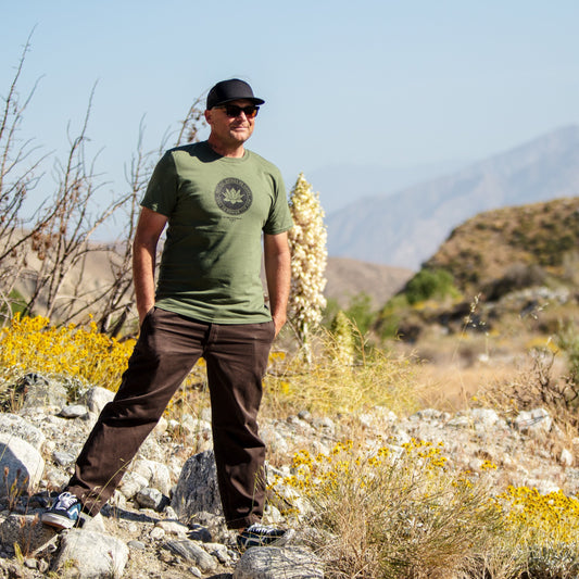 Person in a green Buddhist lotus shirt with the text, May All Beings Be Happy May All Beings Be Free, they are wearing sunglasses standing in a desert landscape with mountains in the background.