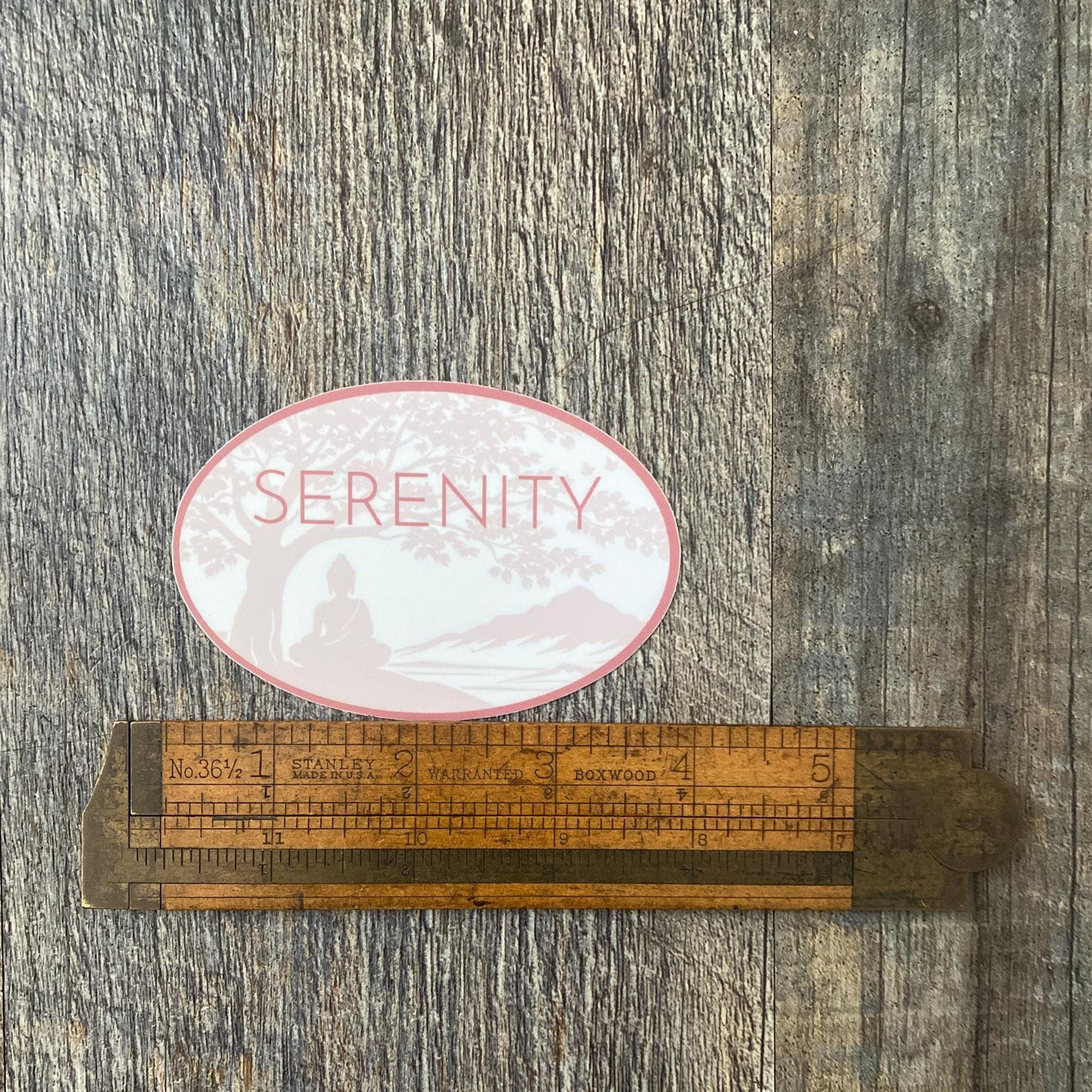 Oval shaped Serenity sticker with a Buddha under a Bodhi Tree. It is being measured by vintage ruler on a wooden surface