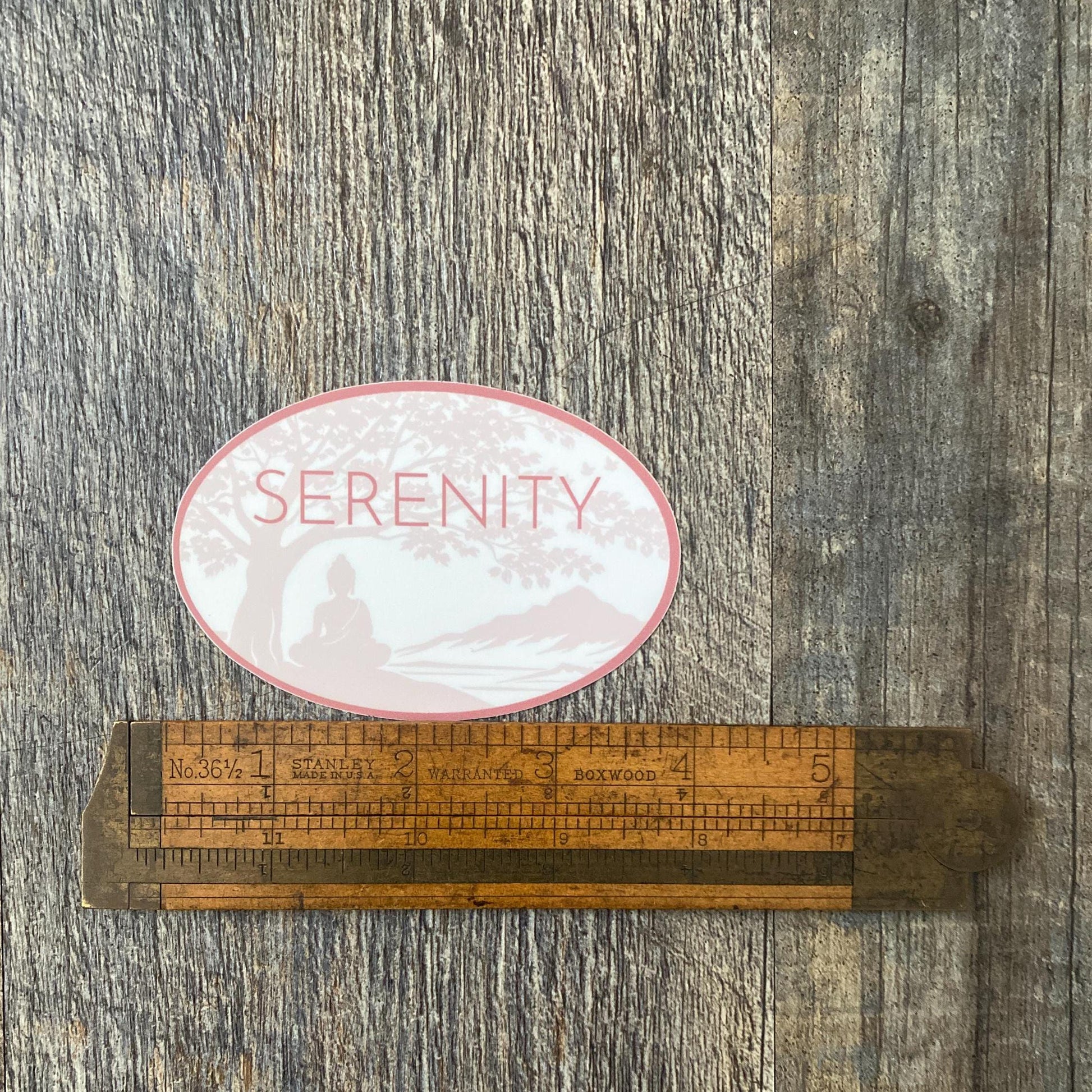 Oval shaped Serenity sticker with a Buddha under a Bodhi Tree. It is being measured by vintage ruler on a wooden surface