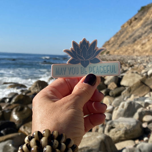 Hand holding a lotus flower sticker with 'May you be peaceful' text on a rocky beach.