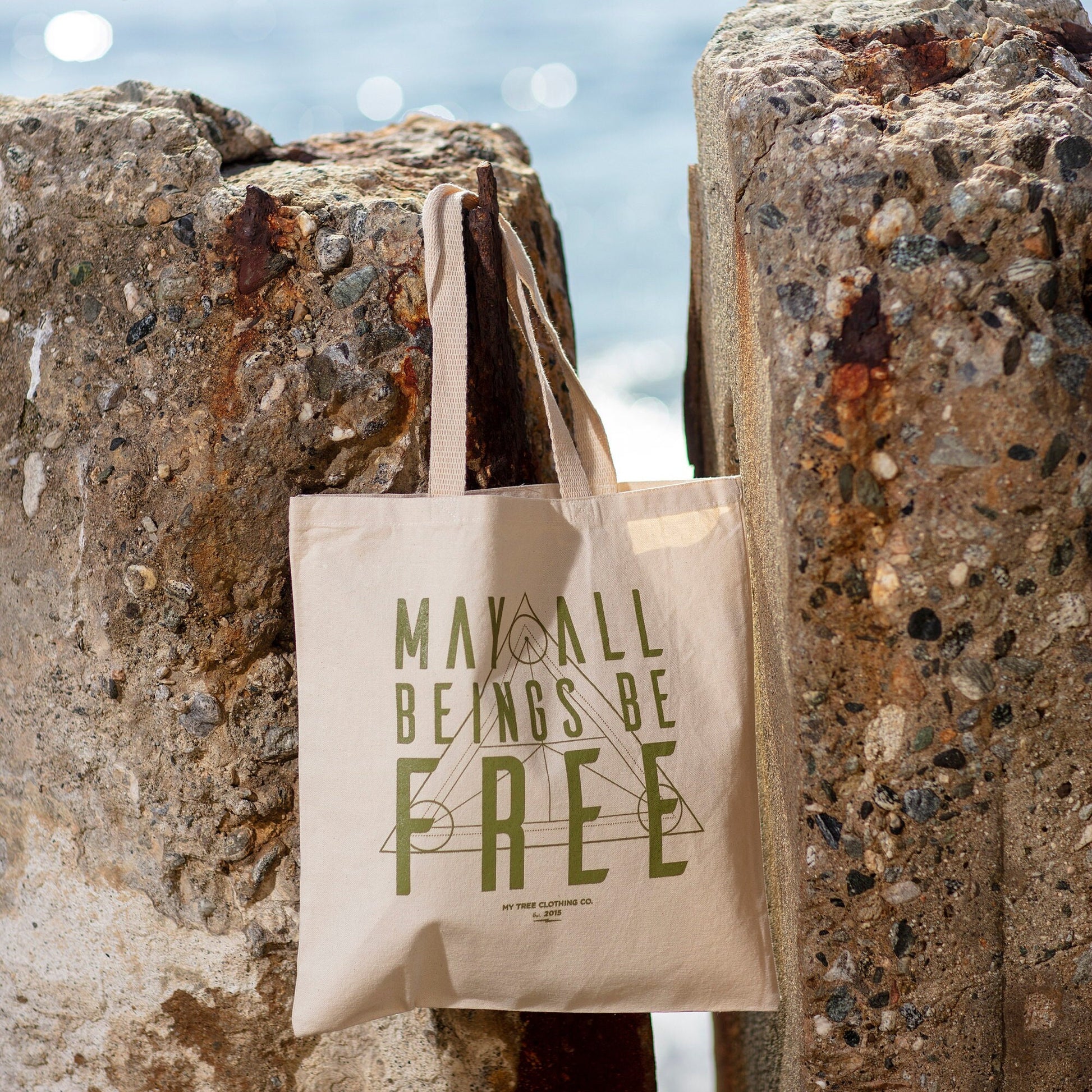 Beige tote bag with 'May All Beings Be Free' text against a textured stone wall.