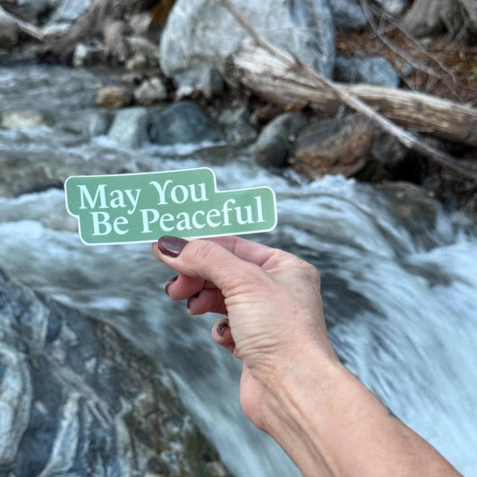 Hand holding a 4" green and white sticker that reads 'May You Be Peaceful' in front of a flowing stream.