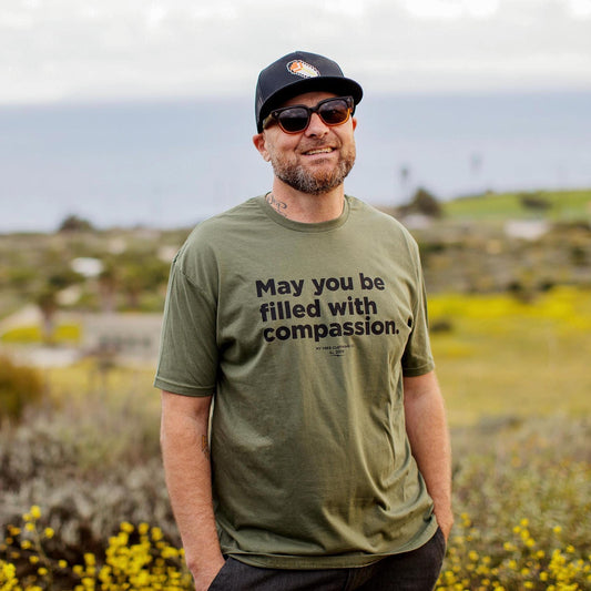 Man wearing a green t-shirt with a message, May You Be Filled With Compassion. They are wearing sunglasses, and a cap in a natural setting.