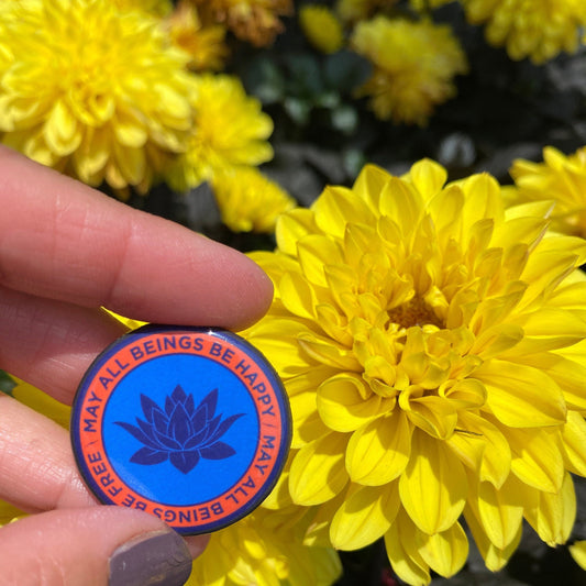 Hand holding a 1.25" circular pin back button with a lotus flower design and text, may all beings be free may all beings be happy.