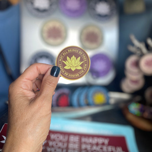 Hand holding a 2" round saffron and gold vinyl sticker with a Buddhist mettā prayer text, may all beings be free, may all beings be happy against a blurred background of similar stickers.