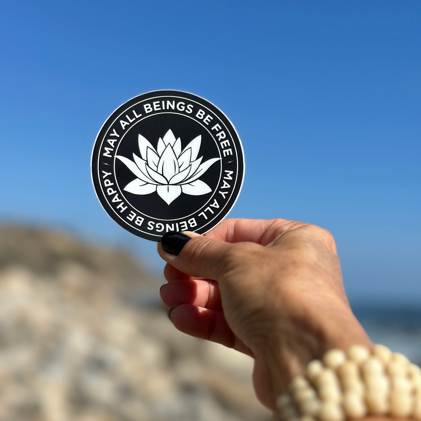 Hand holding a circular sticker with a lotus flower design and the metta prayer against a blue sky.