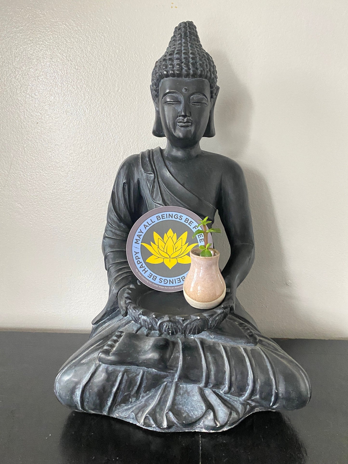 Buddha statue with a 4" round Buddhist sticker with yellow buddhist lotus flower design and text, may all beings be free may all beings be happy. Original design by My Tree Clothing Co.