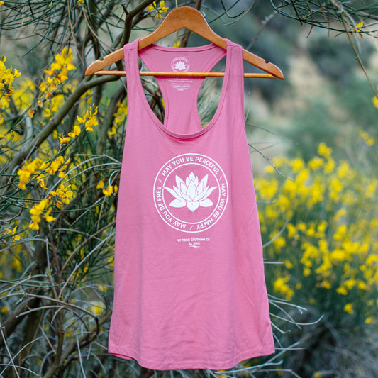 Pink tank top with a white lotus flower design and text that reads, 'may you be peaceful may you be happy may you be free' on a hanger against a natural background with yellow flowers.