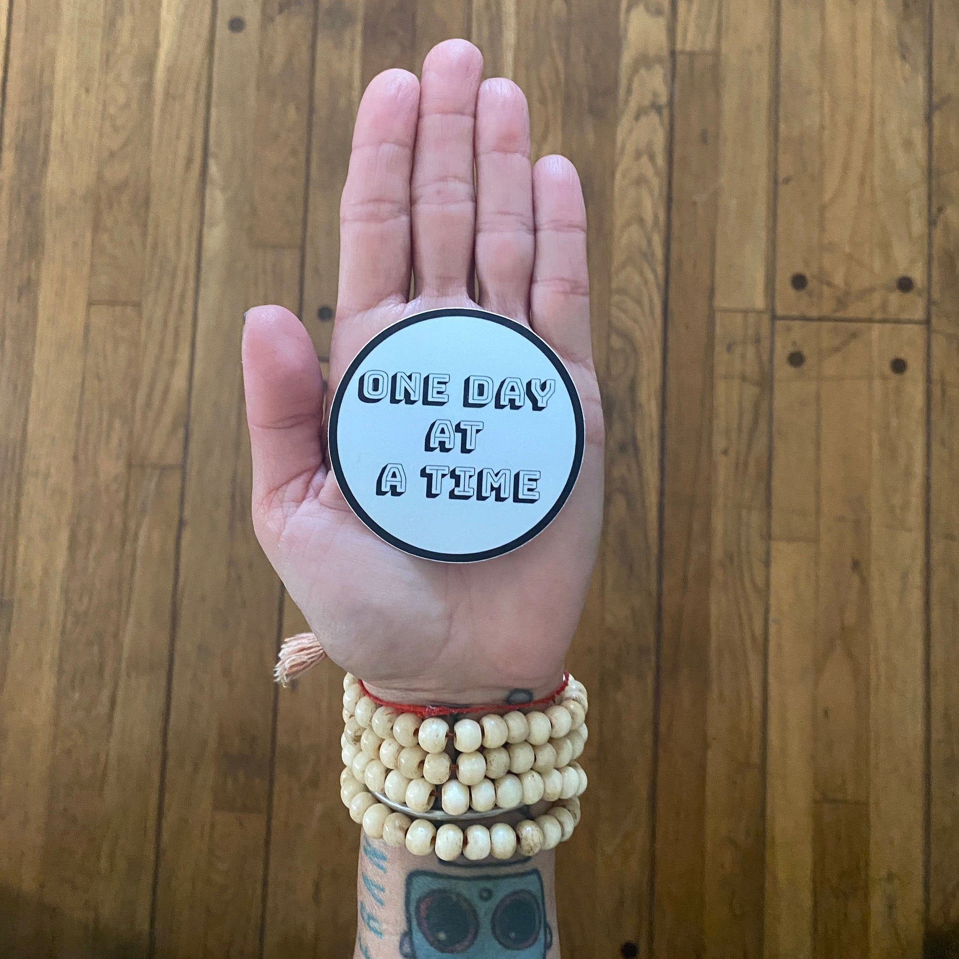 Hand holding a round sticker with 'ONE DAY AT A TIME' text against a wooden background
