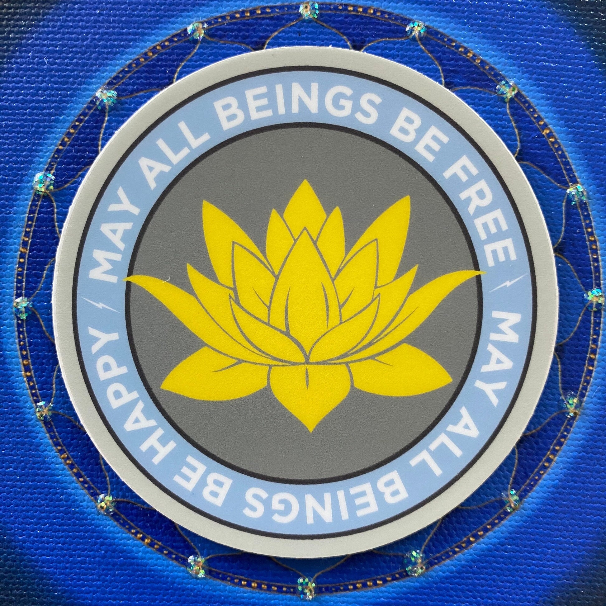 3" circular sticker with a yellow lotus flower and text 'MAY ALL BEINGS BE FREE, HAPPY, HARMONY' on a blue background.