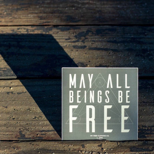 5" square sticker with 'May all beings be free' text with sacred geometry design on a wooden surface.
