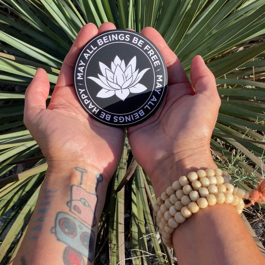 Person holding a 3.5" round black and white magnet with lotus flower design and text, may all beings be free, may all beings be happy, against a natural background.