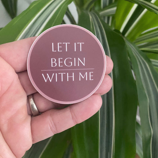 Round pink sticker with the Al-Anon declaration, 'Let It Begin With Me' text held in a hand against a green leafy background.