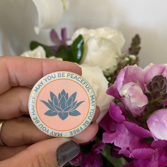 Person holding a circular button with a lotus flower design and text, may you be happy may you be peaceful, may you be free, surrounded by flowers.