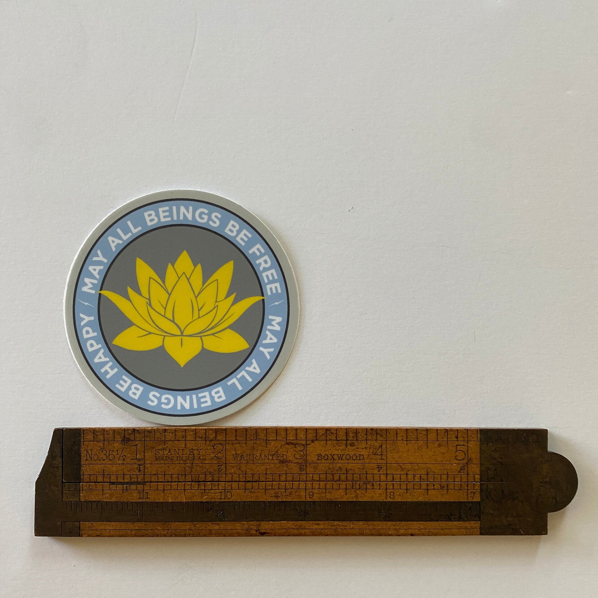 3" circular sticker with a lotus flower design and text, may all beings be free may all beings be happy, next to a wooden ruler for scale.