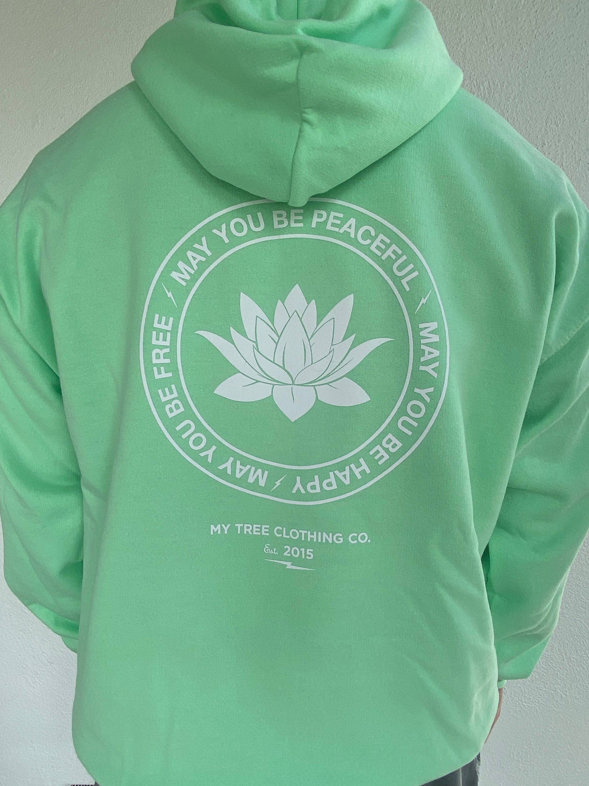 Green hoodie with a white lotus flower design and text may you be peaceful may you be happy may you be free on the back against a plain background.