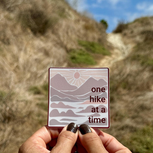 Hand holding a 3" square sticker with 'one hike at a time' in neutral colors with mountains and a sunset against a natural background.