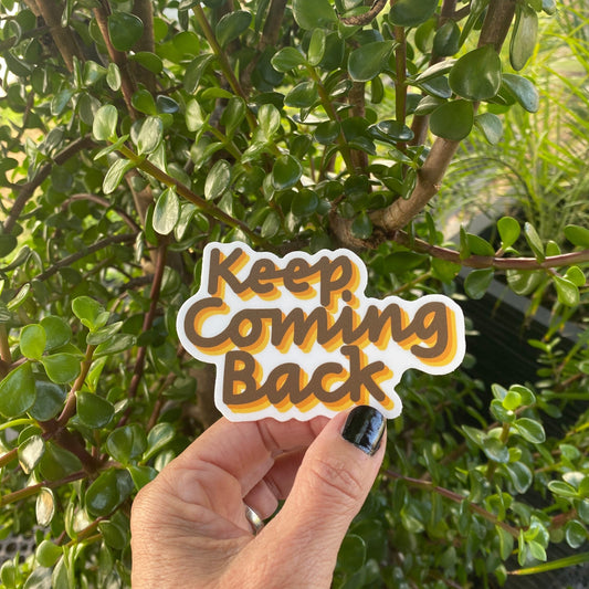 Hand holding a 'Keep Coming Back' sticker in front of green foliage.