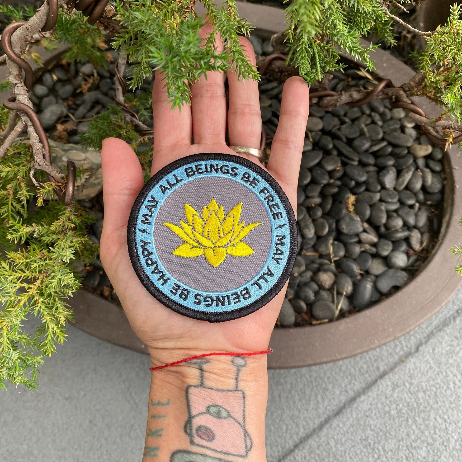 Hand holding 3" round embroidered Buddhist patch with a golden lotus flower design and the text, may all beings be free may all beings be happy, against a natural background.