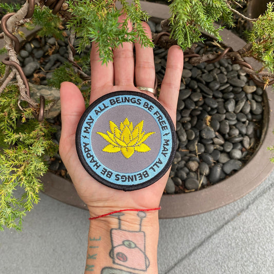 Hand holding 3" round embroidered Buddhist patch with a golden lotus flower design and the text, may all beings be free may all beings be happy, against a natural background.