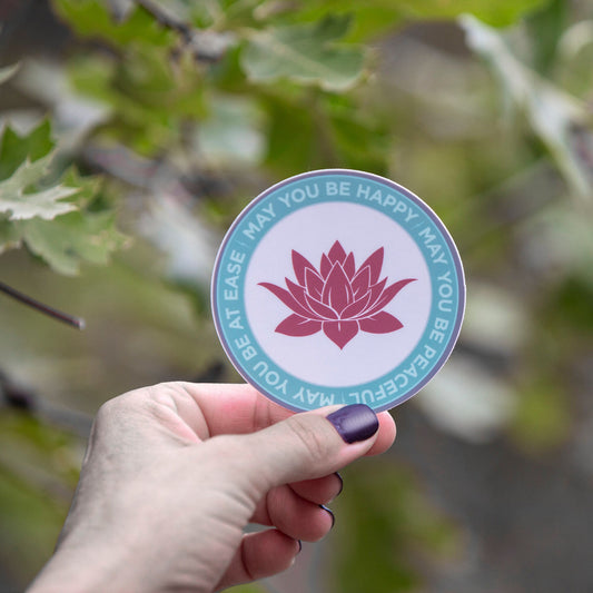 Hand holding a circular sticker with a lotus flower design that reads, "may you be happy may you be peaceful may you be at ease" against a blurred natural background.