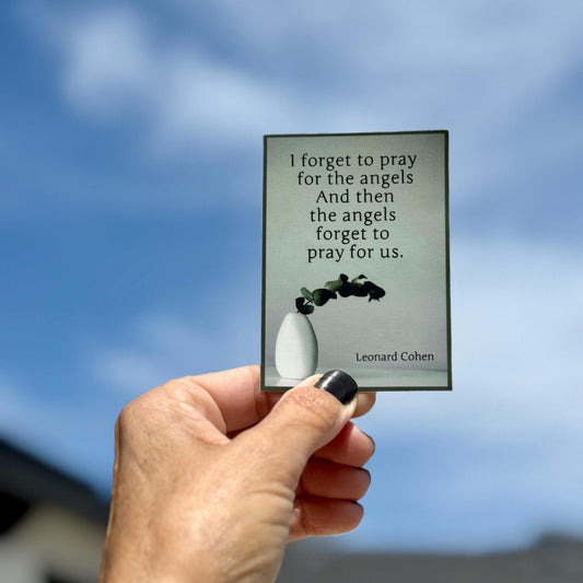 Hand holding a rectangular sticker with a Leonard Cohen quote, 'I forget to pray for the angels and then the angels forget to pray for us.' It is being held against a blue sky.