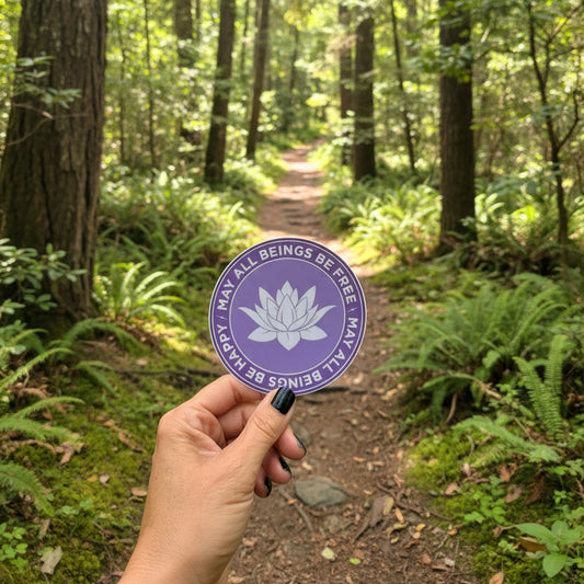 Purple sticker with text, may all beings be free-may all beings be happy with a lotus flower held by a hand against a natural background.